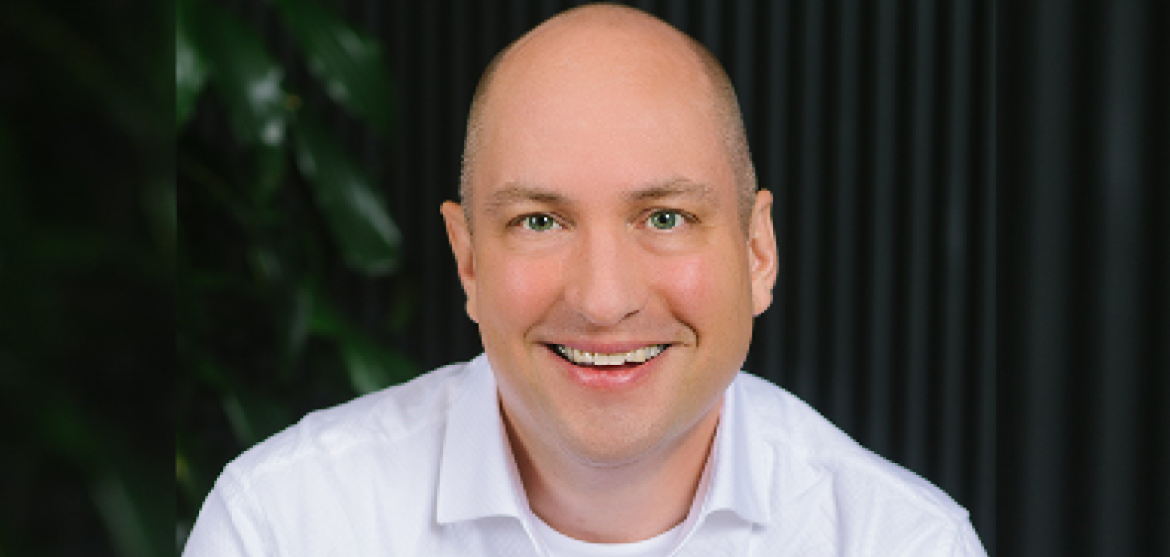 A bald white man wearing a white shirt sits before a dark background with a plant to his left