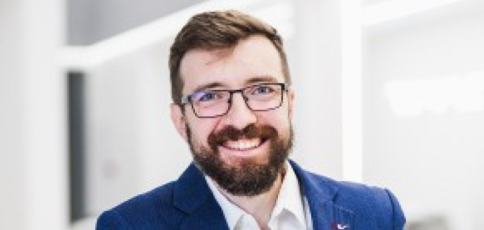A white man with brown hair, glasses and a beard wearing a blue suit