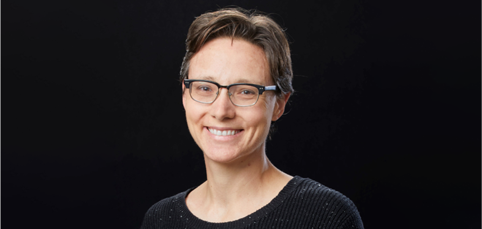 A woman with short brown hair and glasses wears a dark jumper and sits in front of a dark background