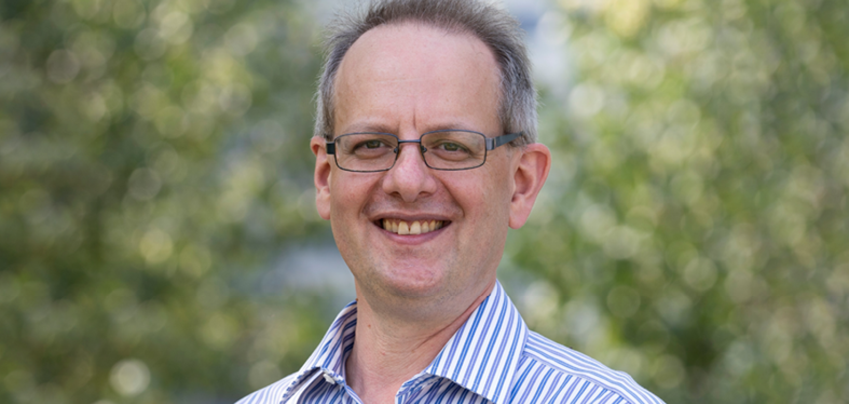 A white man with glasses and wearing a striped shirt, stands before a leafy background