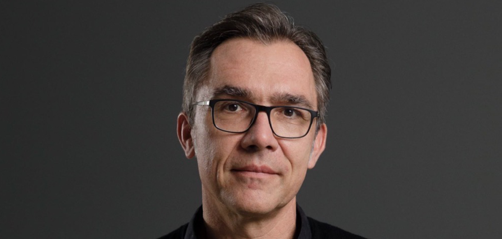 A white man with glasses and wearing a black shirt stands in front of a grey background