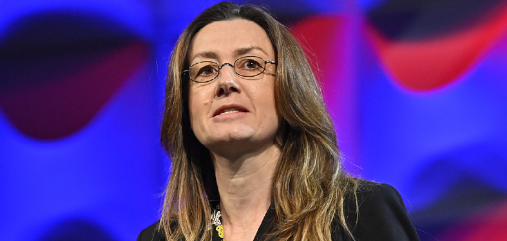 A woman with long brown hair stands before a multicoloured background