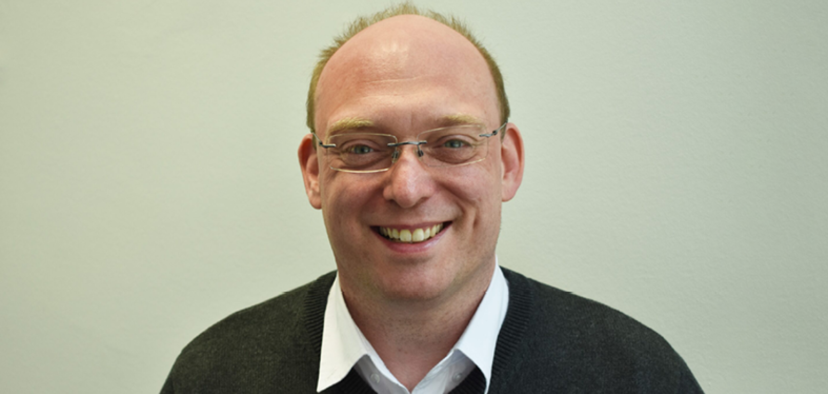 A white man with glasses and wearing a white shirt and jumper sits in fronto og a green background