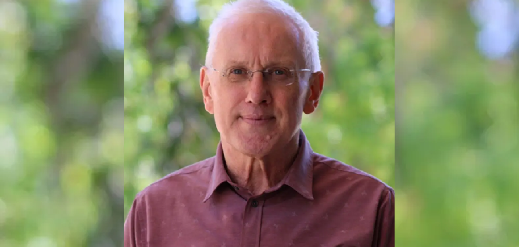 A man with white hair wearing a red shirt stands in front of a leafy background