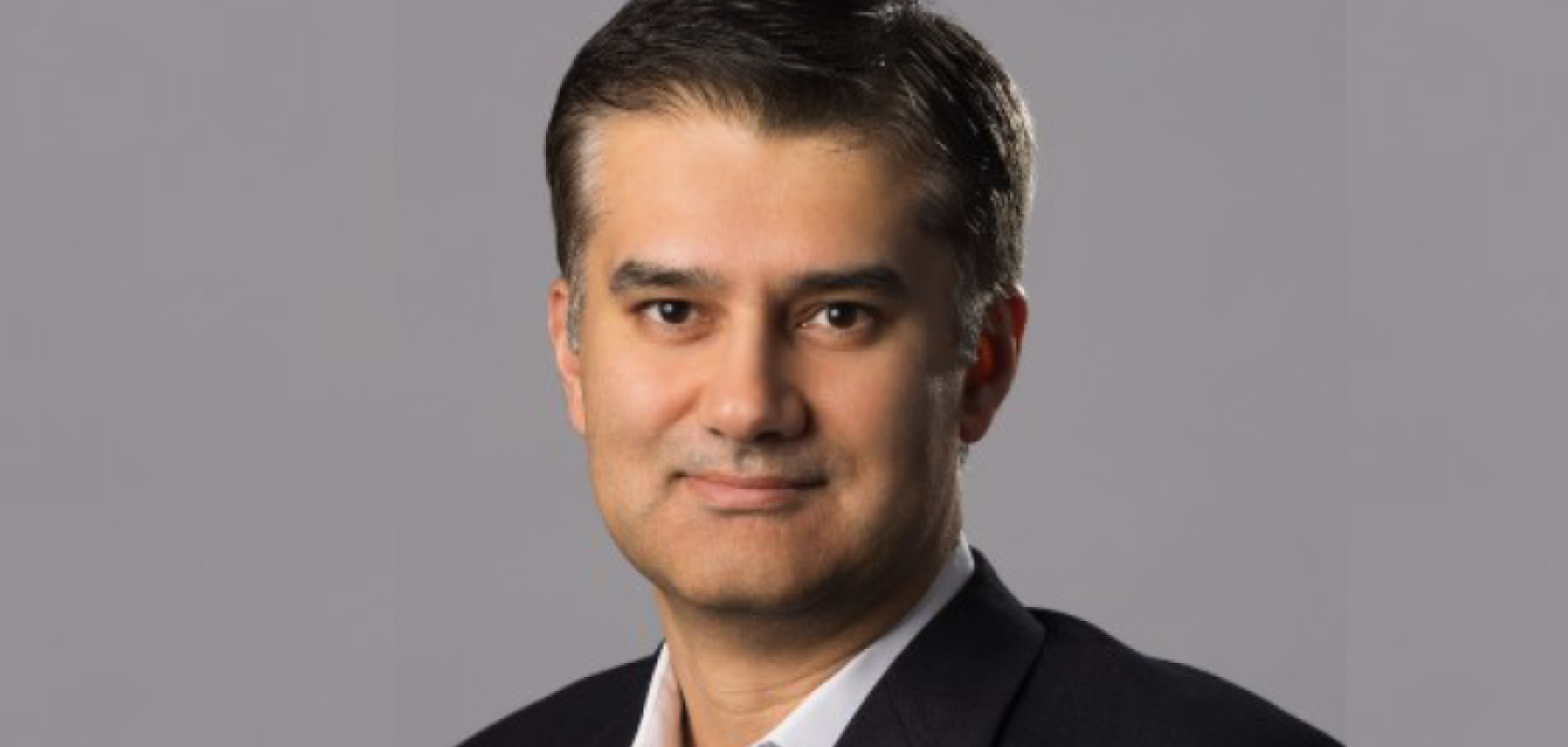 A man with dark brown hair and a dark suit stands in front of a grey background