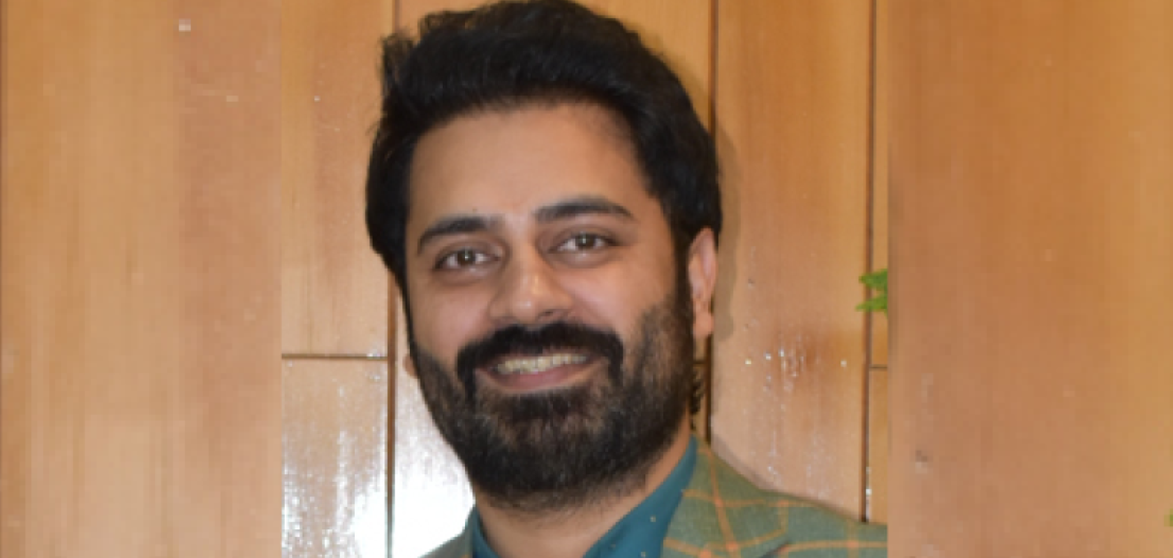 An Asian man withdark hair and a beard is wearing a green check suit and standing before a woodpanelled wall