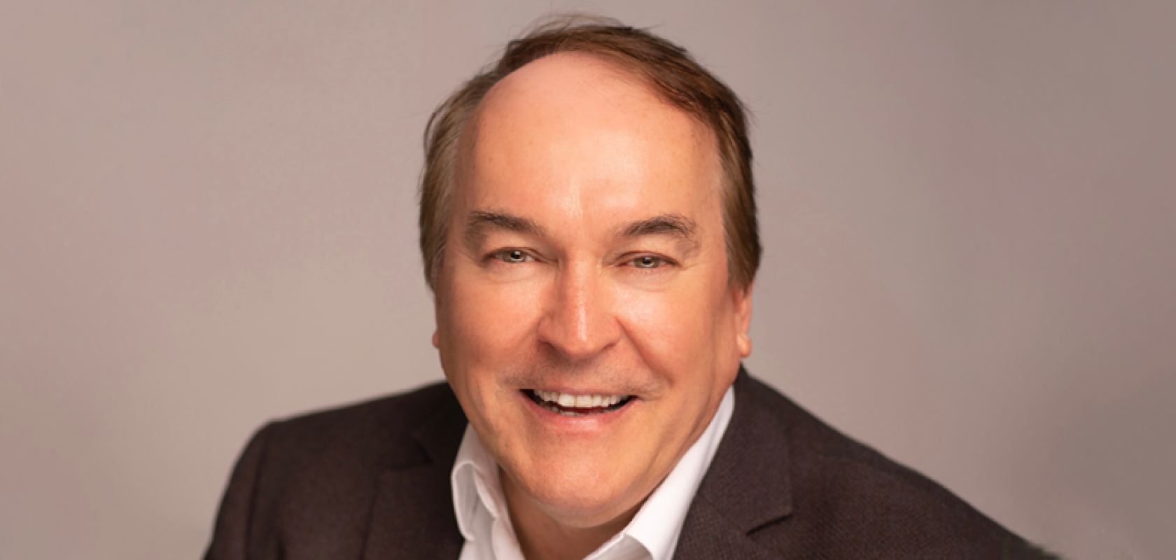 A white, clean shaven man wearing a brown suit sits in front of a beige background
