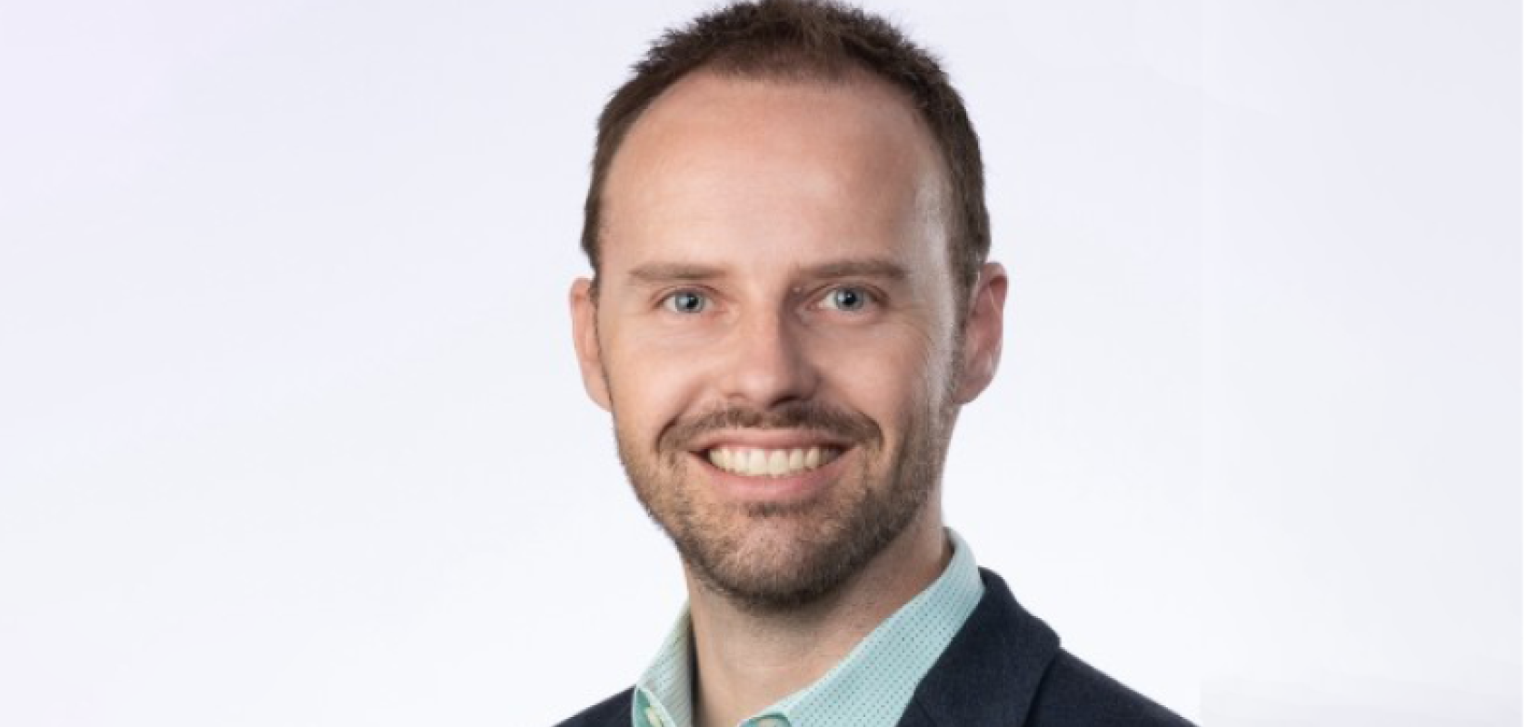 A white man with a light beard smiles at the camera. He is wearing a blue suit and a green shirt 