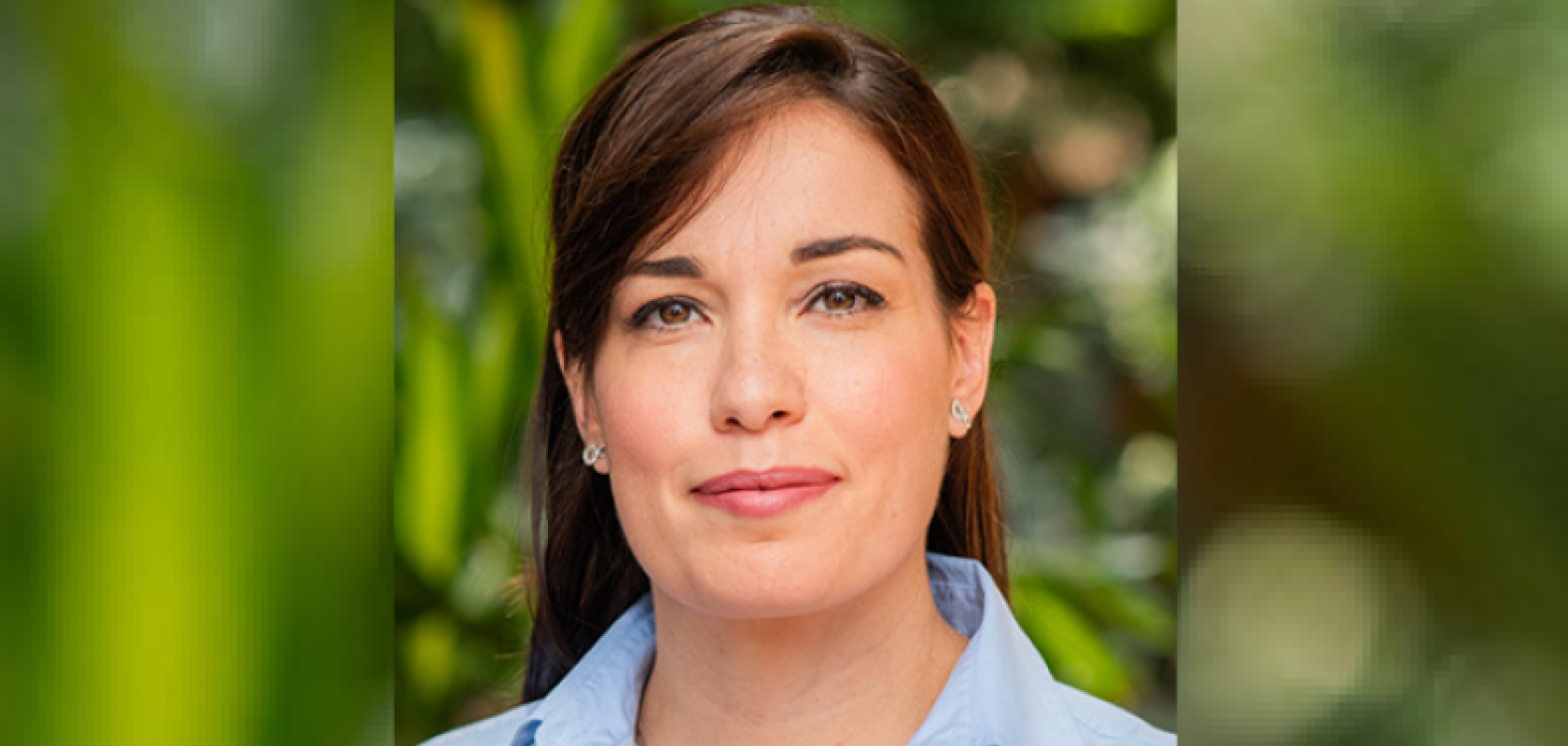 A white woman with long brown hair and brown eyes wears a blue shirt in front of some green foliage