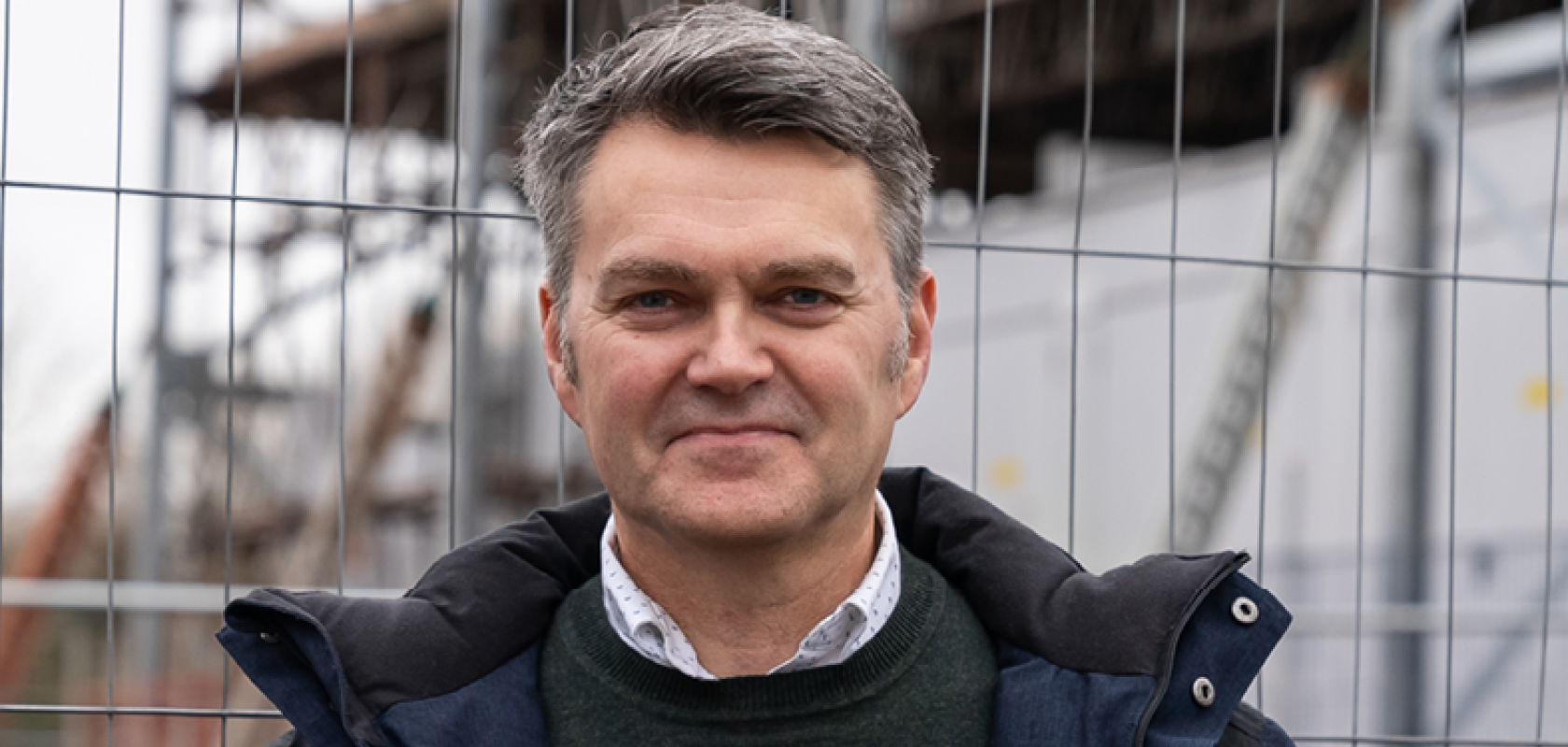 A white man with greying hair wears a blue raincoat in front of a chainlink fence