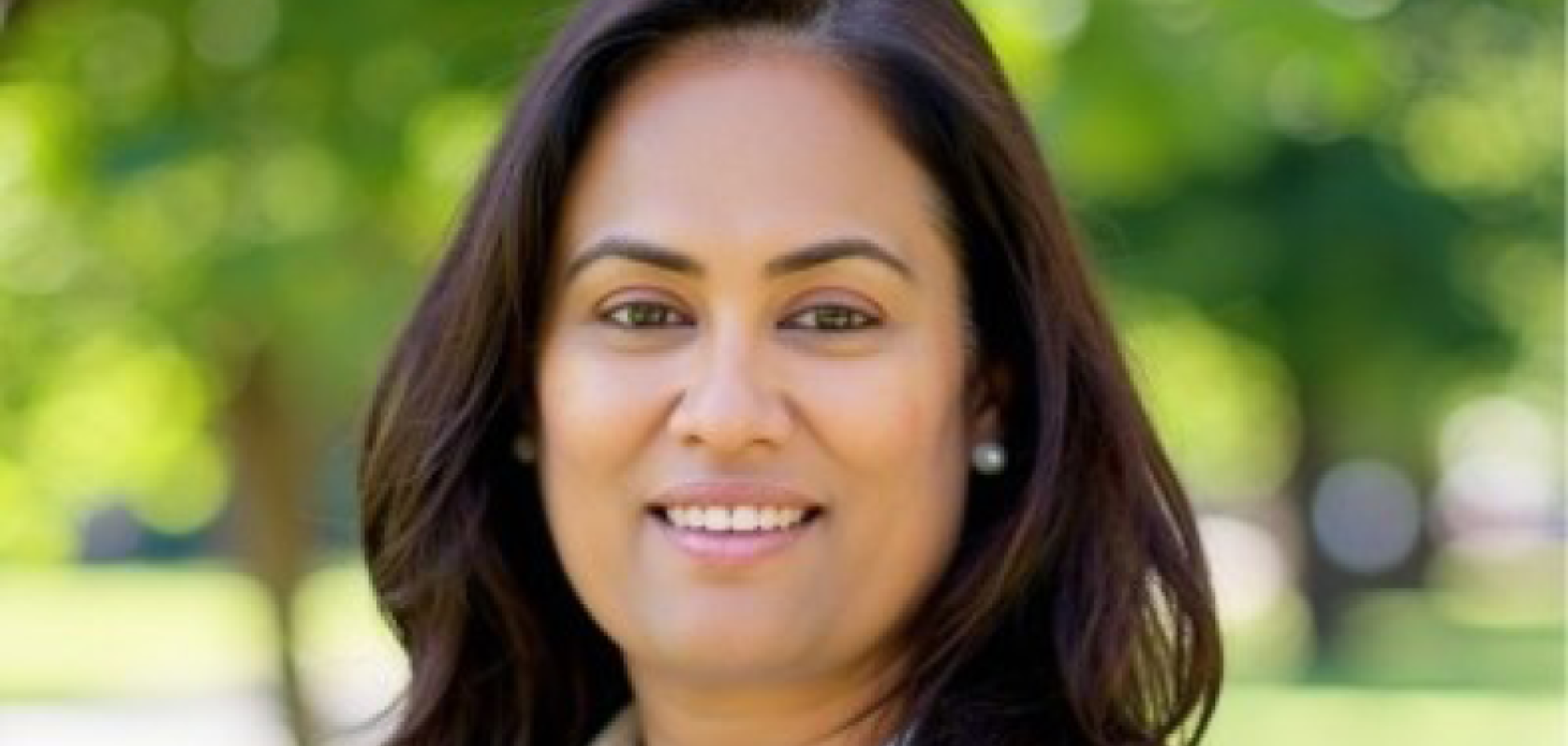 A woman with dark brown hair and brown eyes wears a white shirt. She is standing in front of a leafy park