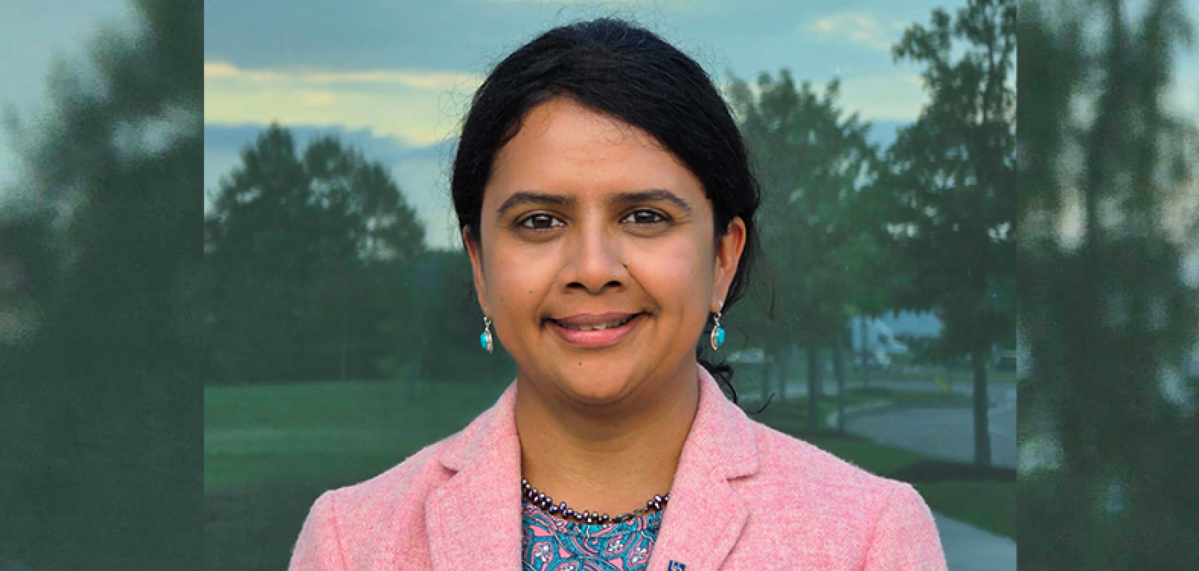 An Asian woman with dark brown hair and a pink jacket stands in front of a leafy background