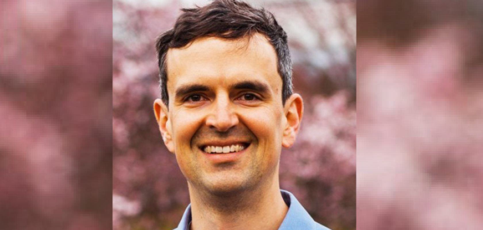A white man with short brown hair. He is smiing and standing in front of some red trees