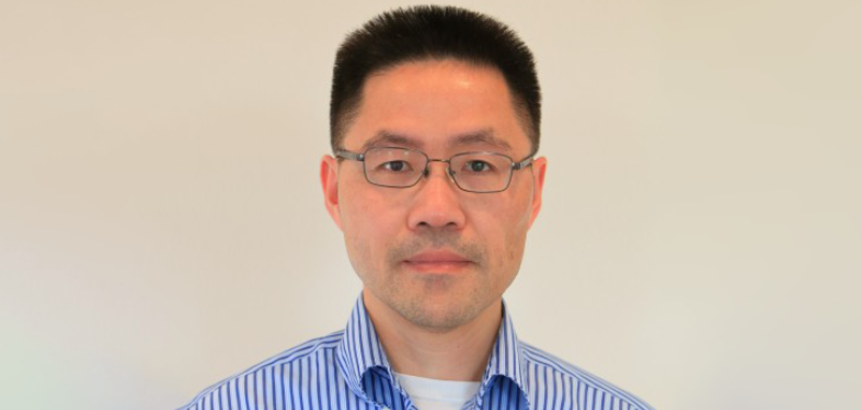 An Asian man with glasses and wearing a blue and white striped shirt stands before a beige background