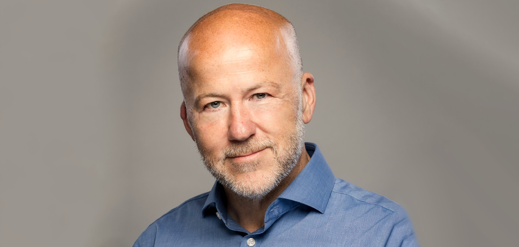 A white, bald man with stubble wearing a blue shirt against a beige background